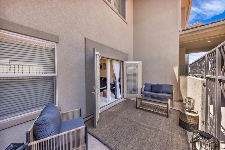 Bright, open-air patio space featuring neutral-toned textured flooring, built-in seating with blue cushioning, mirrored closet doors with louvered ventilation, roller shades on adjacent windows, and abundant natural light from multiple glass sliding doors
