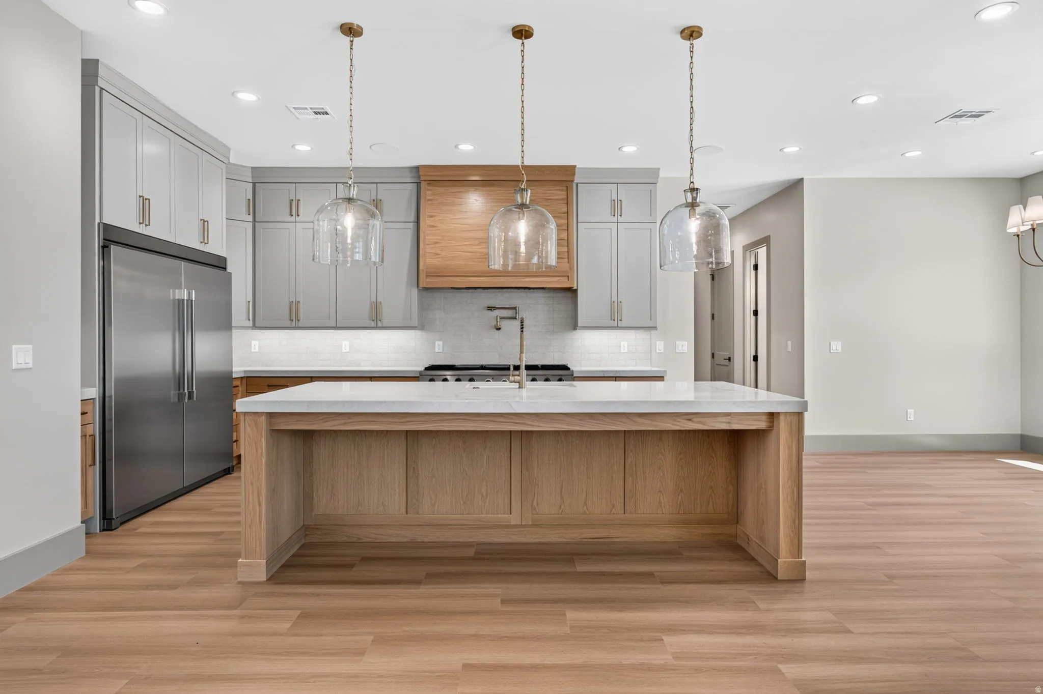 Kitchen featuring a large central island with a light-colored countertop, light wood-finish flooring, stainless steel refrigerator, gray cabinetry, and brass-finish pendant lighting