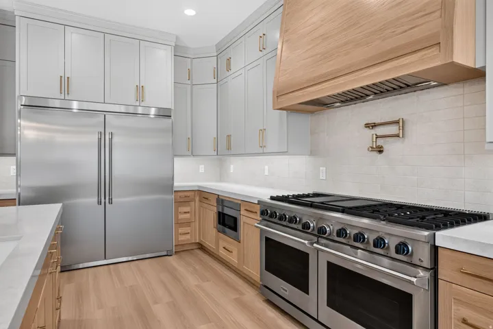 Gourmet kitchen featuring a built-in stainless steel refrigerator, professional-grade range with double ovens, wood-finish flooring, light grey upper cabinetry, and a light wood range hood