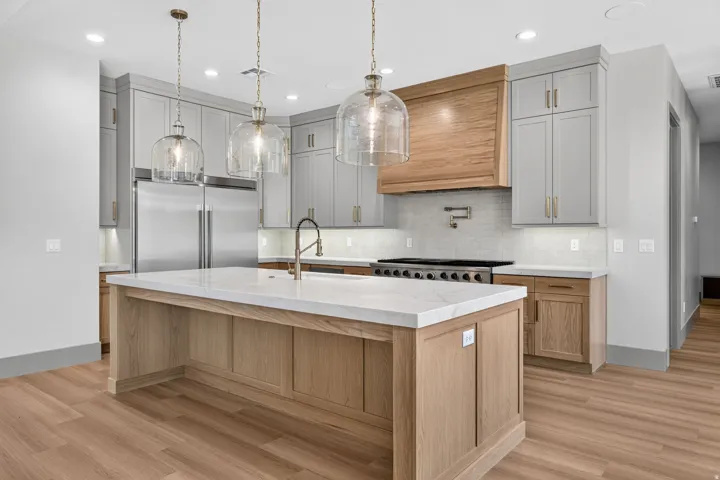 Modern kitchen featuring a large central island with a light-toned countertop, wood-finish flooring, two-tone cabinetry, and stainless steel appliances