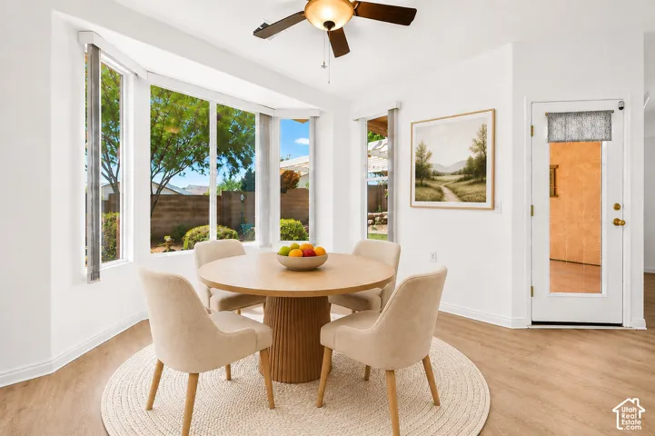 Bright dining area showcasing expansive multi-pane windows framing outdoor scenery, wood-finish flooring, a round pedestal table base, upholstered dining chairs, ceiling fan with brass-finish fixture, and a framed landscape artwork on the accent wall