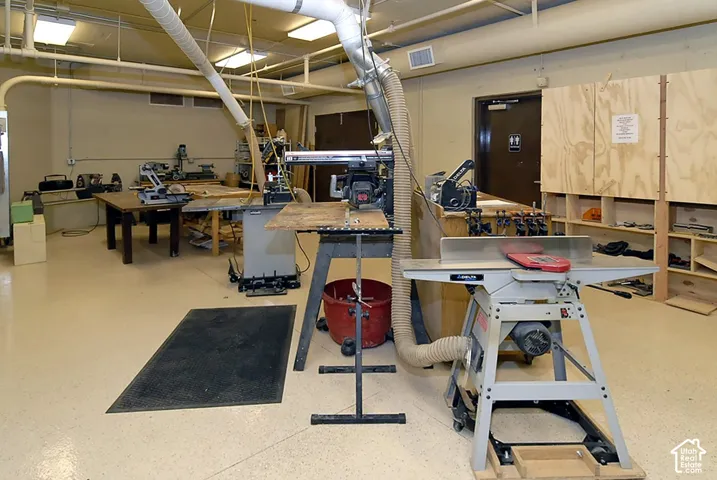 Spacious workshop area featuring light-toned flooring, exposed overhead ductwork, ample fluorescent lighting, and built-in plywood shelving units
