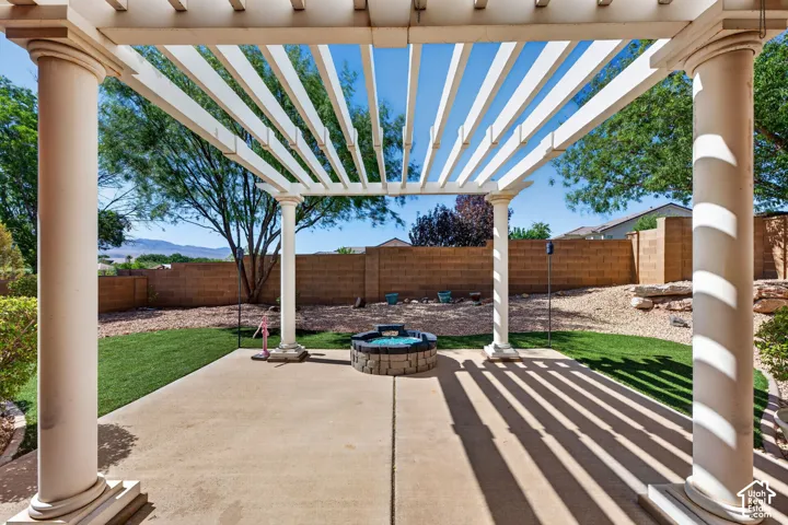 Expansive patio featuring a white pergola with classic columns, a concrete surface, and a built-in stone fire pit, all overlooking a landscaped yard with green turf and a block privacy wall