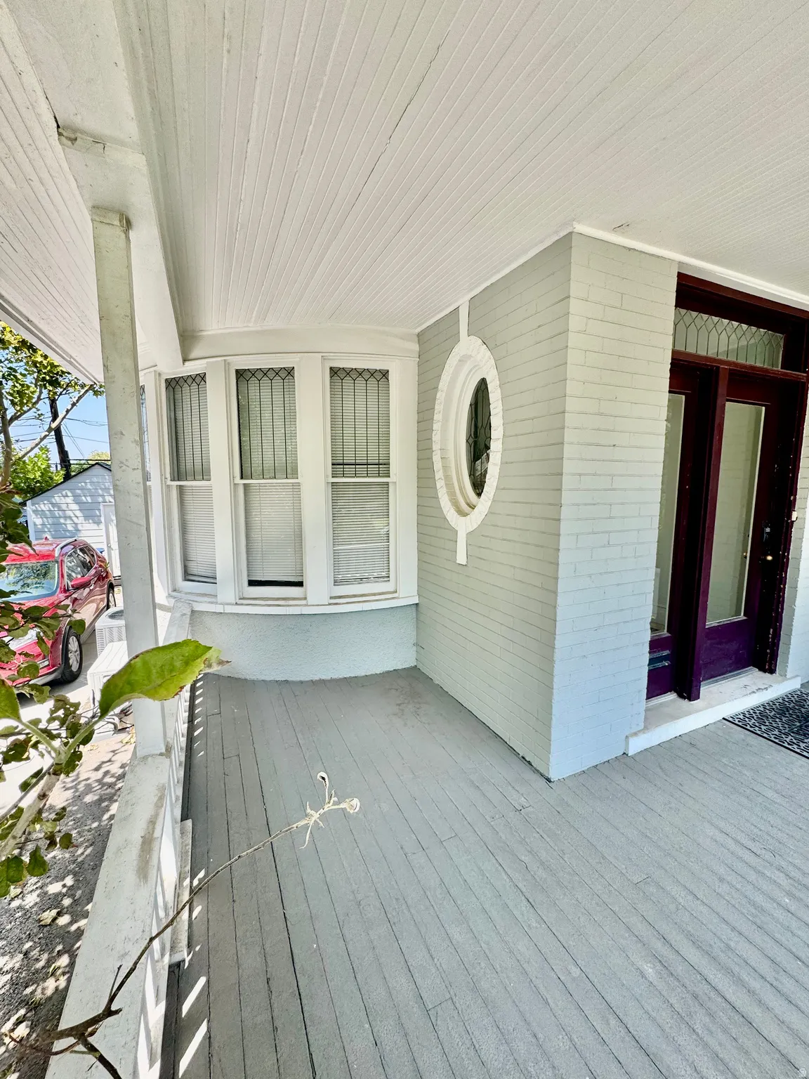 Spacious covered porch with plank flooring, a curved bay window, decorative oval window, and a dark-stained wood entry door with glass panels