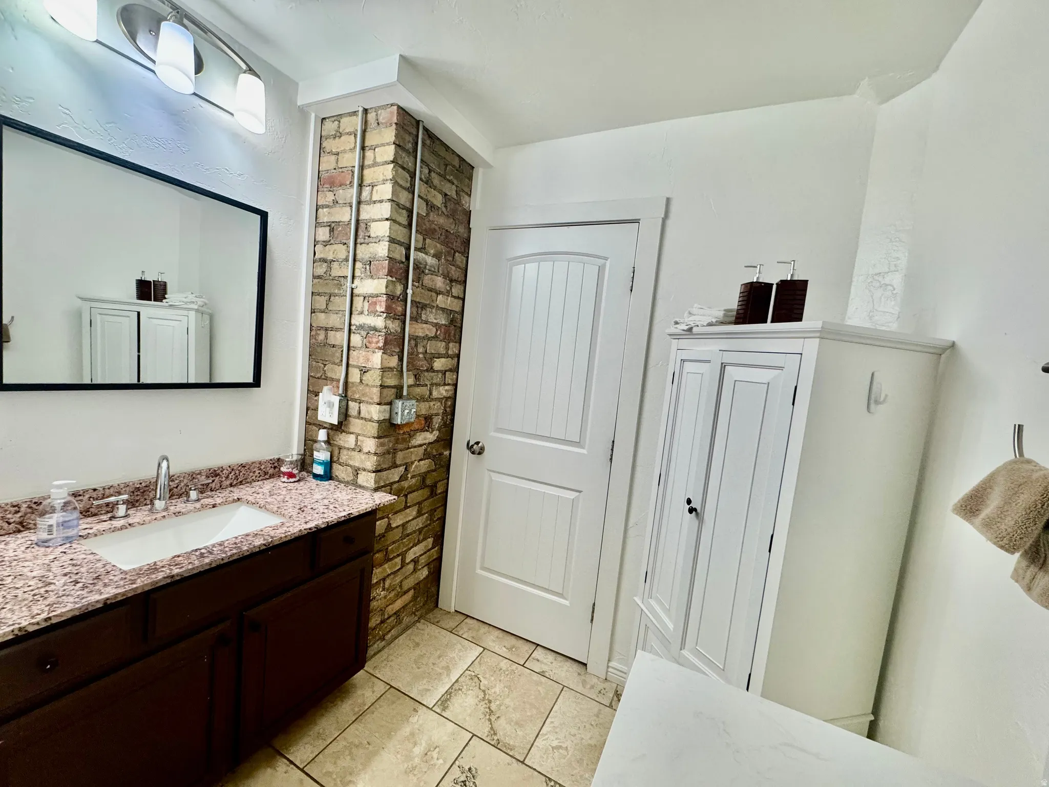 Bathroom featuring a dark wood vanity with a drop-in sink, a granite-finish countertop, a rectangular mirror, and a brick accent wall with exposed conduit