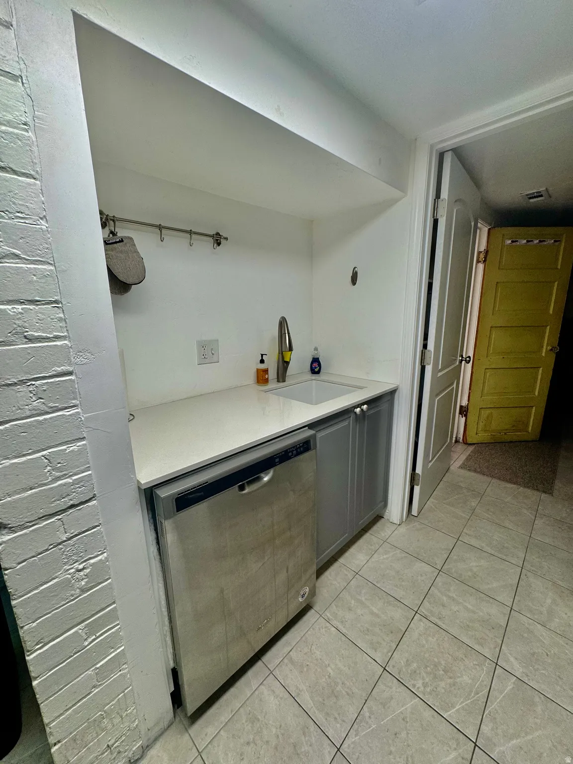 Integrated sink and stainless steel dishwasher set into a white countertop, complemented by gray base cabinetry and tile flooring
