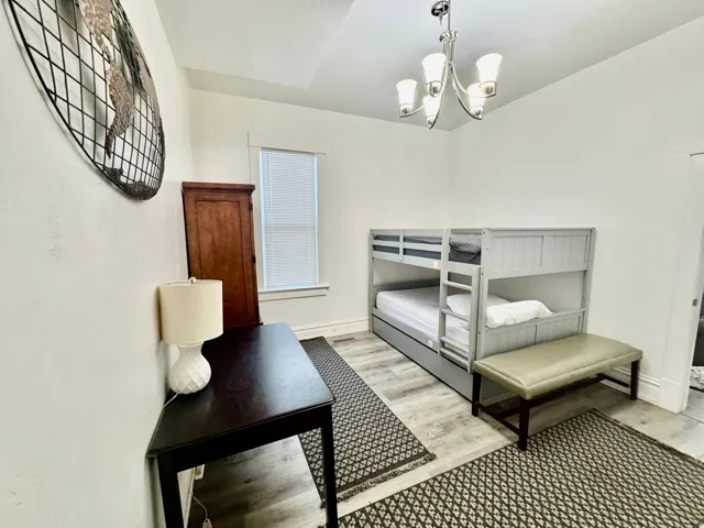 Spacious room featuring light-toned wood-finish flooring, a contemporary chandelier, and a prominent window with blinds