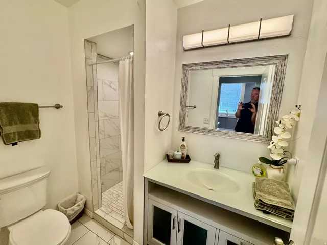 Bathroom featuring a shower with white and gray tile surround, a single vanity with a white countertop and oval sink, and a framed mirror with an overhead light fixture
