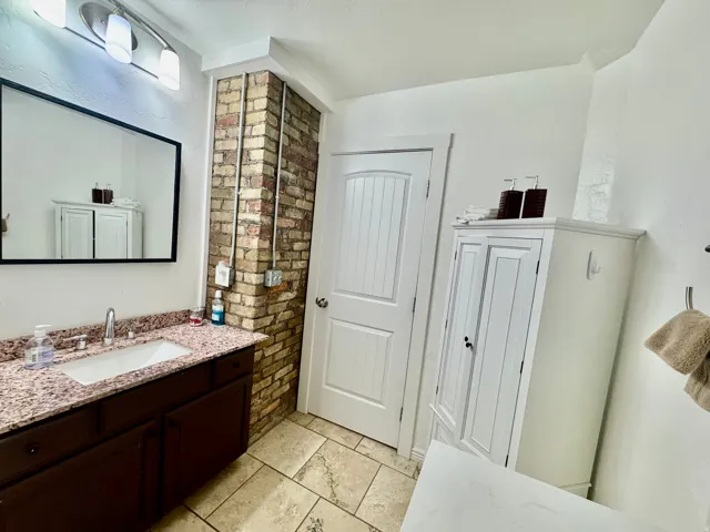 Bathroom featuring a dark wood vanity with a drop-in sink, a granite-finish countertop, a rectangular mirror, and a brick accent wall with exposed conduit