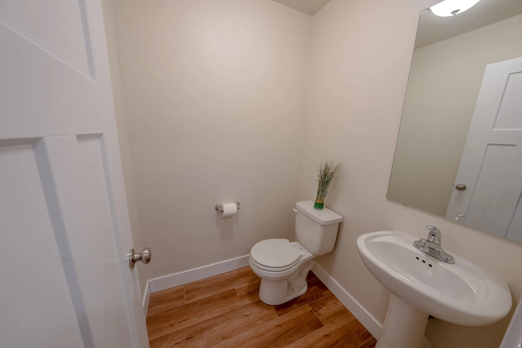 Pedestal sink with chrome faucet, wall-mounted mirror, wood-finish flooring, and white paneled door
