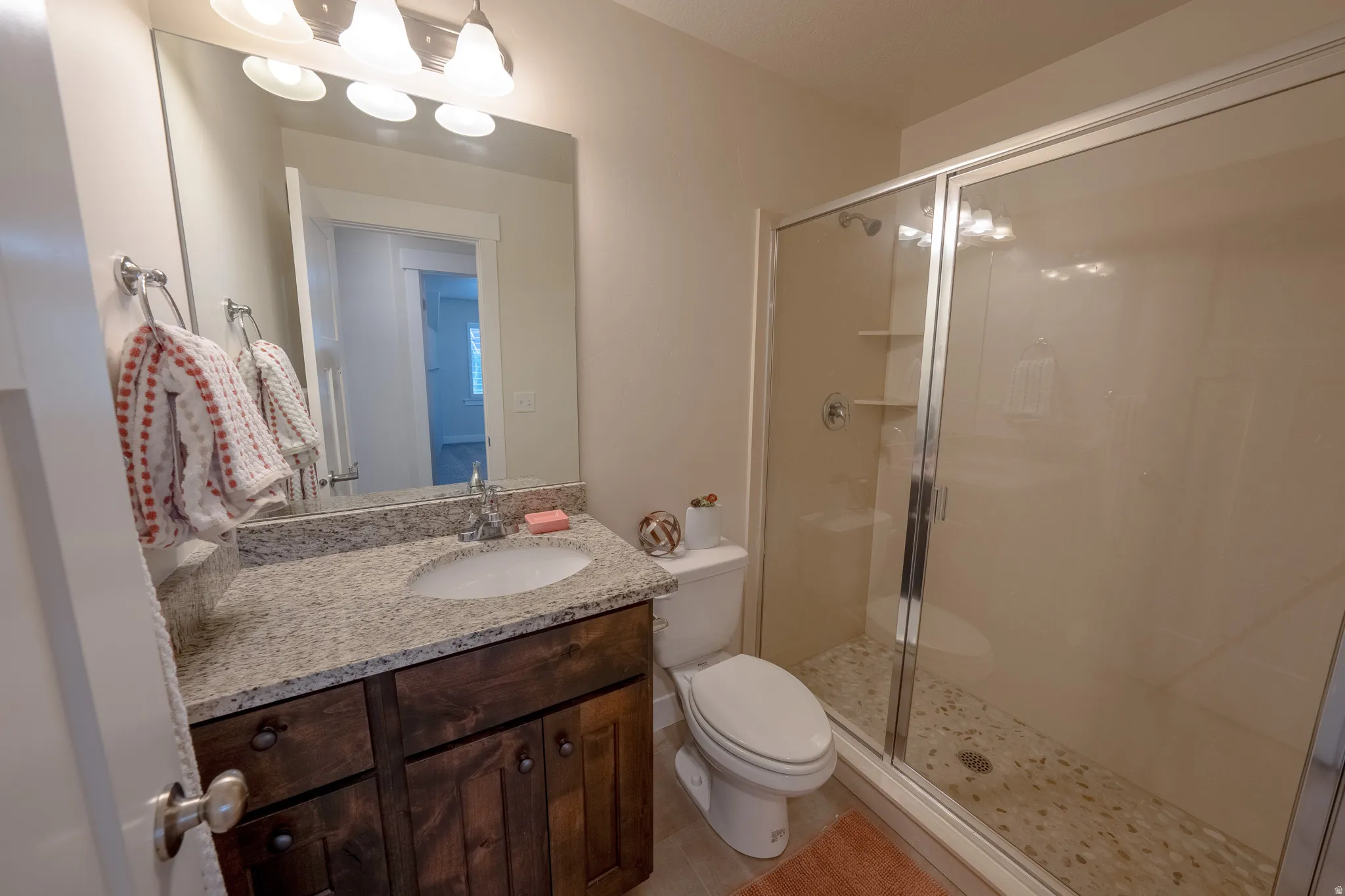 Bathroom featuring a wood-finish vanity with a single basin and stone-finish countertop, a mirror, and a glass-enclosed shower with a pebble-style floor