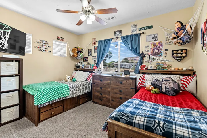 Textured carpeting, window with blinds, ceiling fan, warm neutral wall color, and wood-finish furnishings