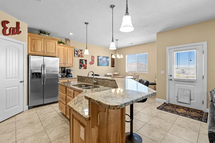 Spacious kitchen featuring stainless steel side-by-side refrigerator, granite-topped island with seating, pendant lighting, wood-finish cabinetry, and tile flooring