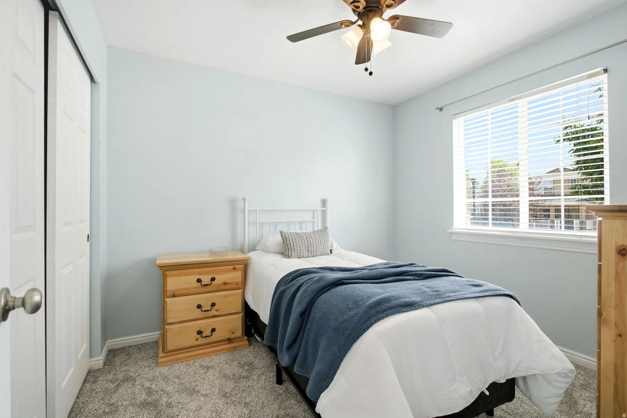 Light blue walls featuring a ceiling fan with integrated lighting, a broad window with white blinds, and gray patterned carpet