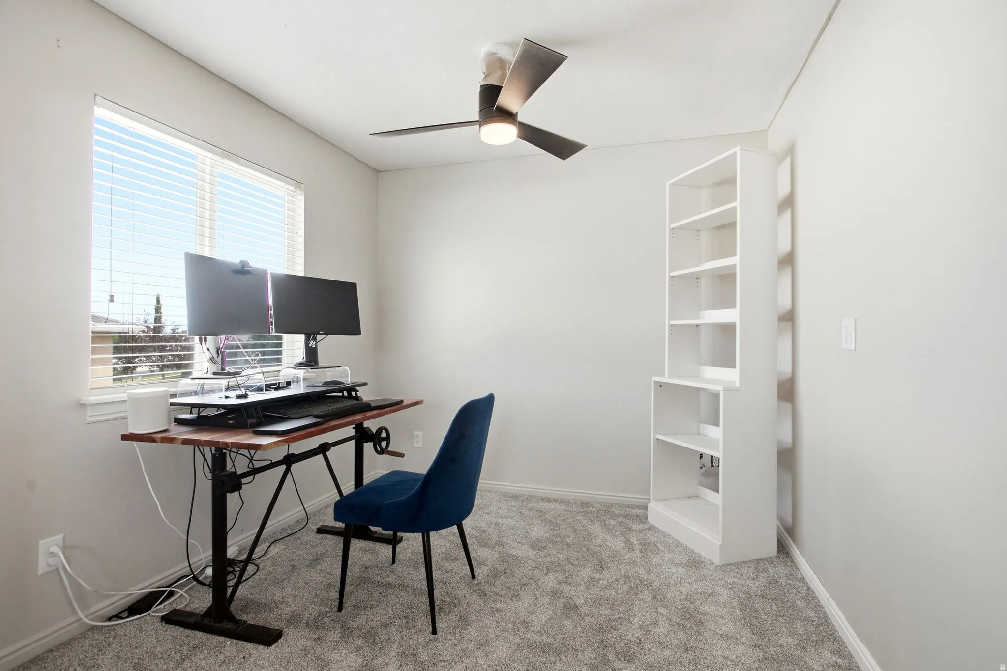 Primary suite sitting room being used as an office with Window with horizontal blinds, overhead ceiling fan with light, light gray wall paint, and gray carpet flooring