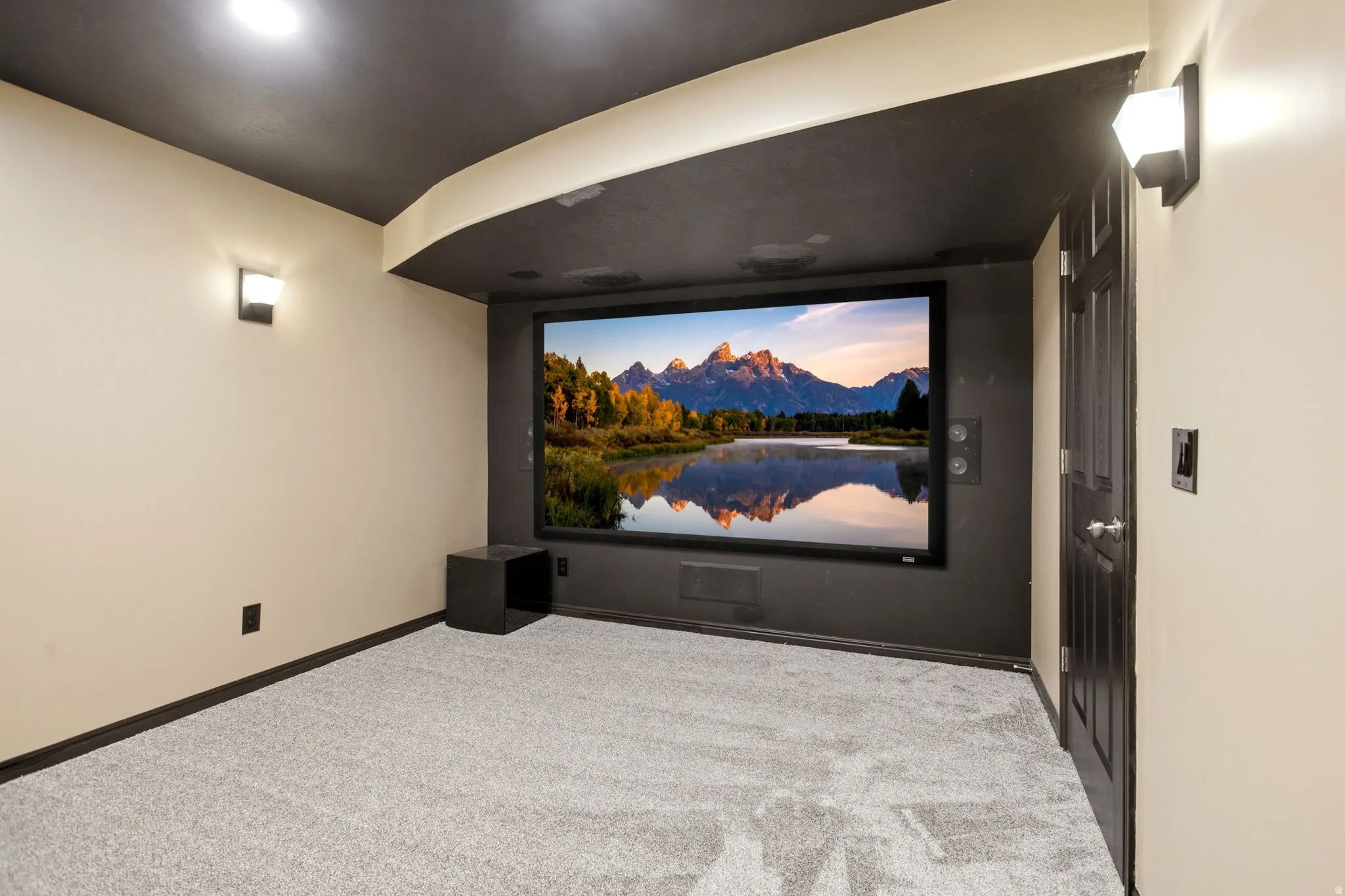 Integrated projection screen, wall-mounted sconce lighting, recessed ceiling lighting, light-colored carpeting, and dark-paneled door