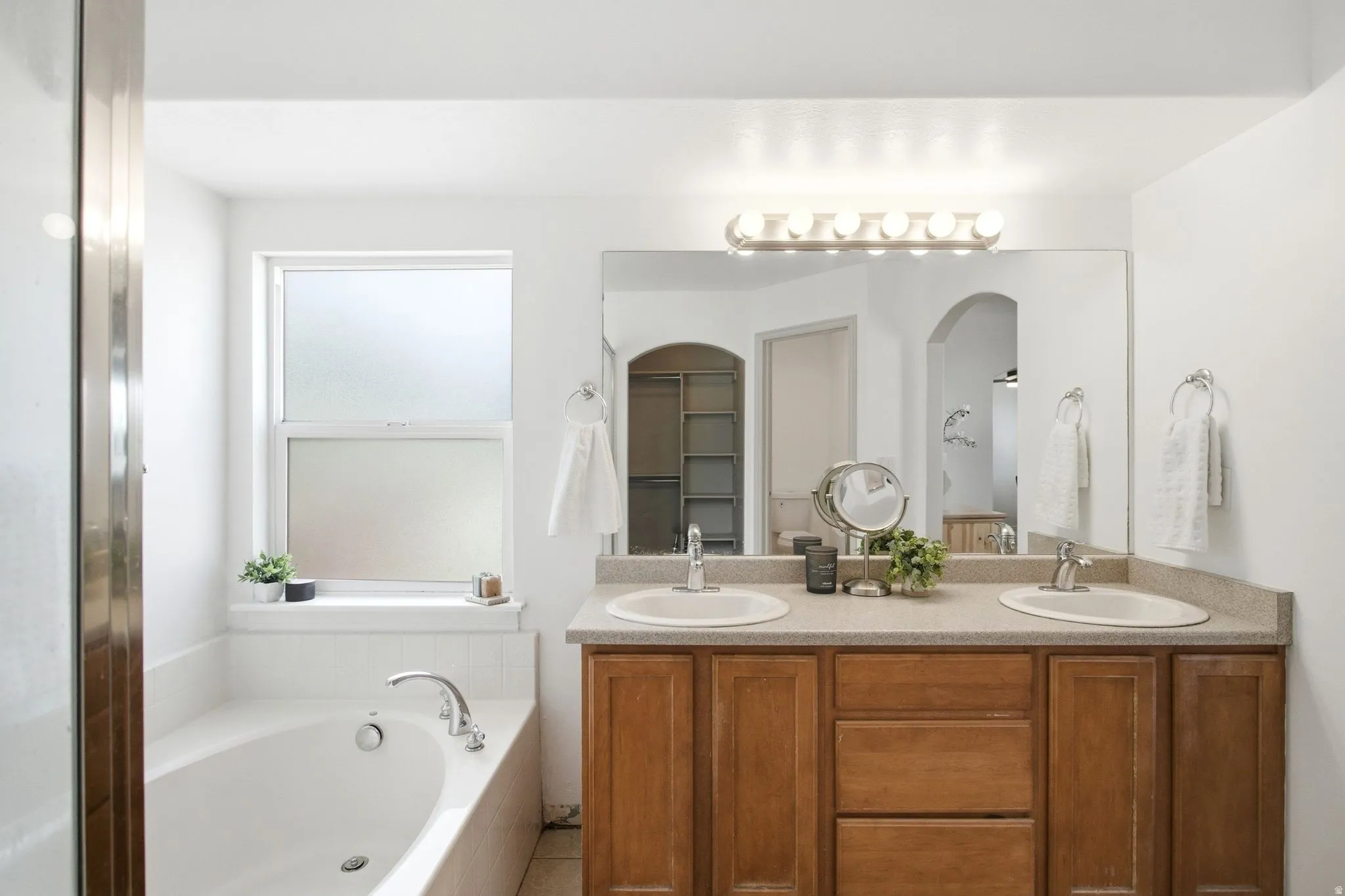 Primary suite Dual vanity with integrated sinks, wood-finish cabinetry, built-in bathtub, frosted window, and large wall-mounted mirror