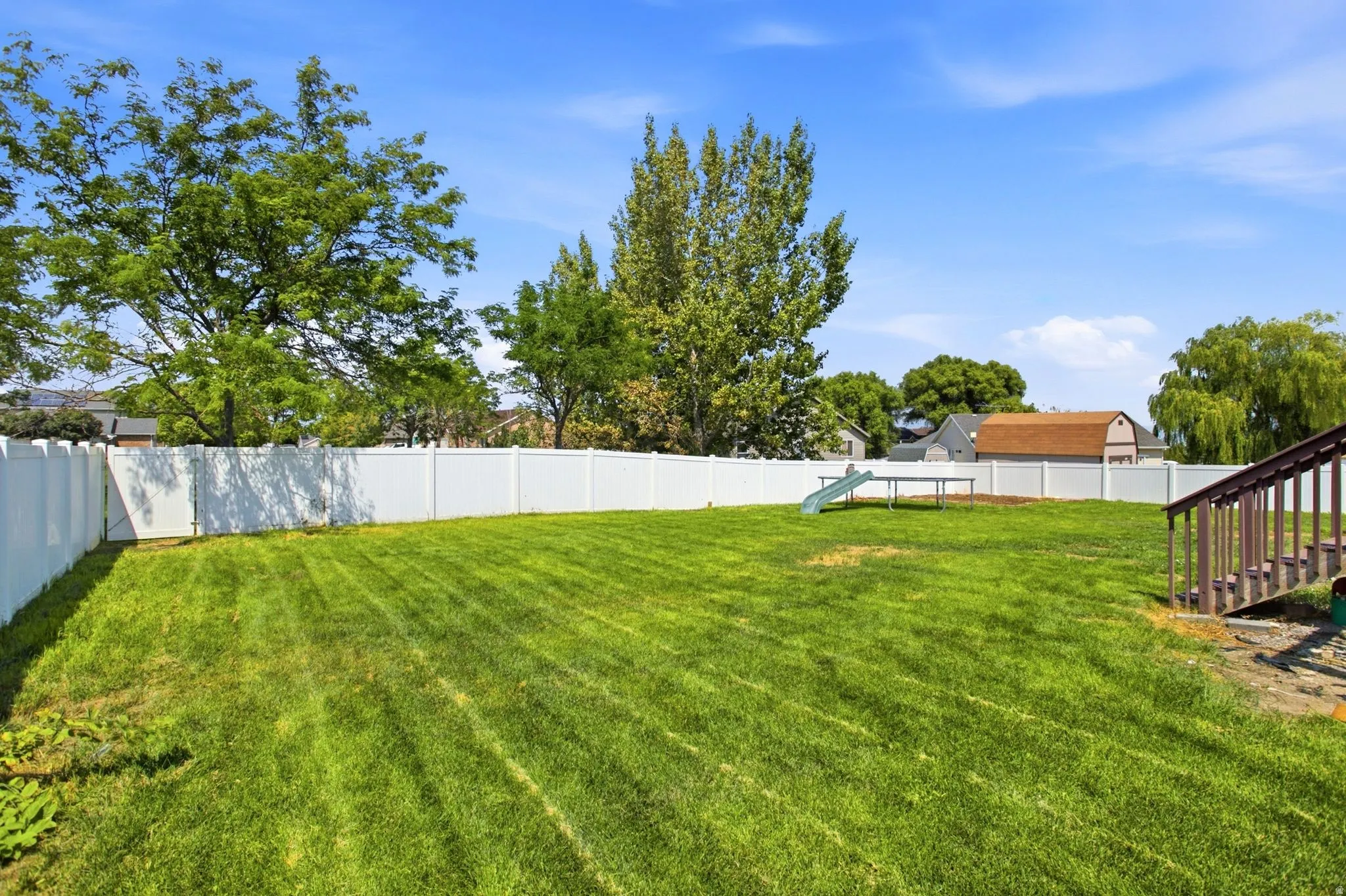 Expansive fenced backyard featuring a manicured lawn, mature trees, and wooden deck stairs