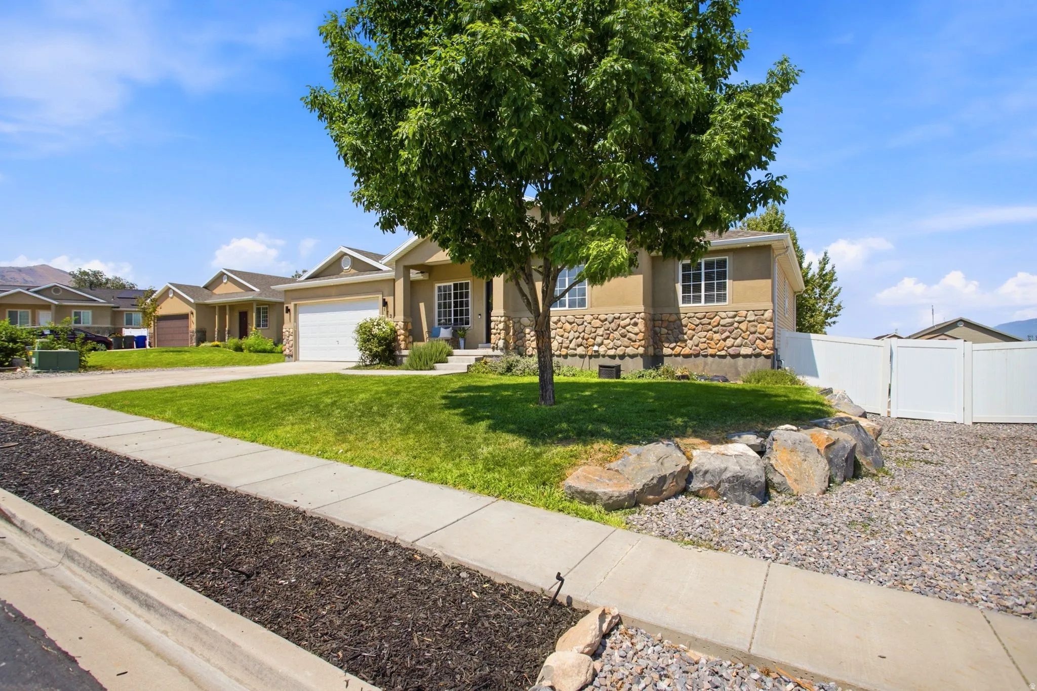 Stucco exterior with stone accenting, integrated two-car garage, manicured lawn, concrete walkway, and white vinyl fencing