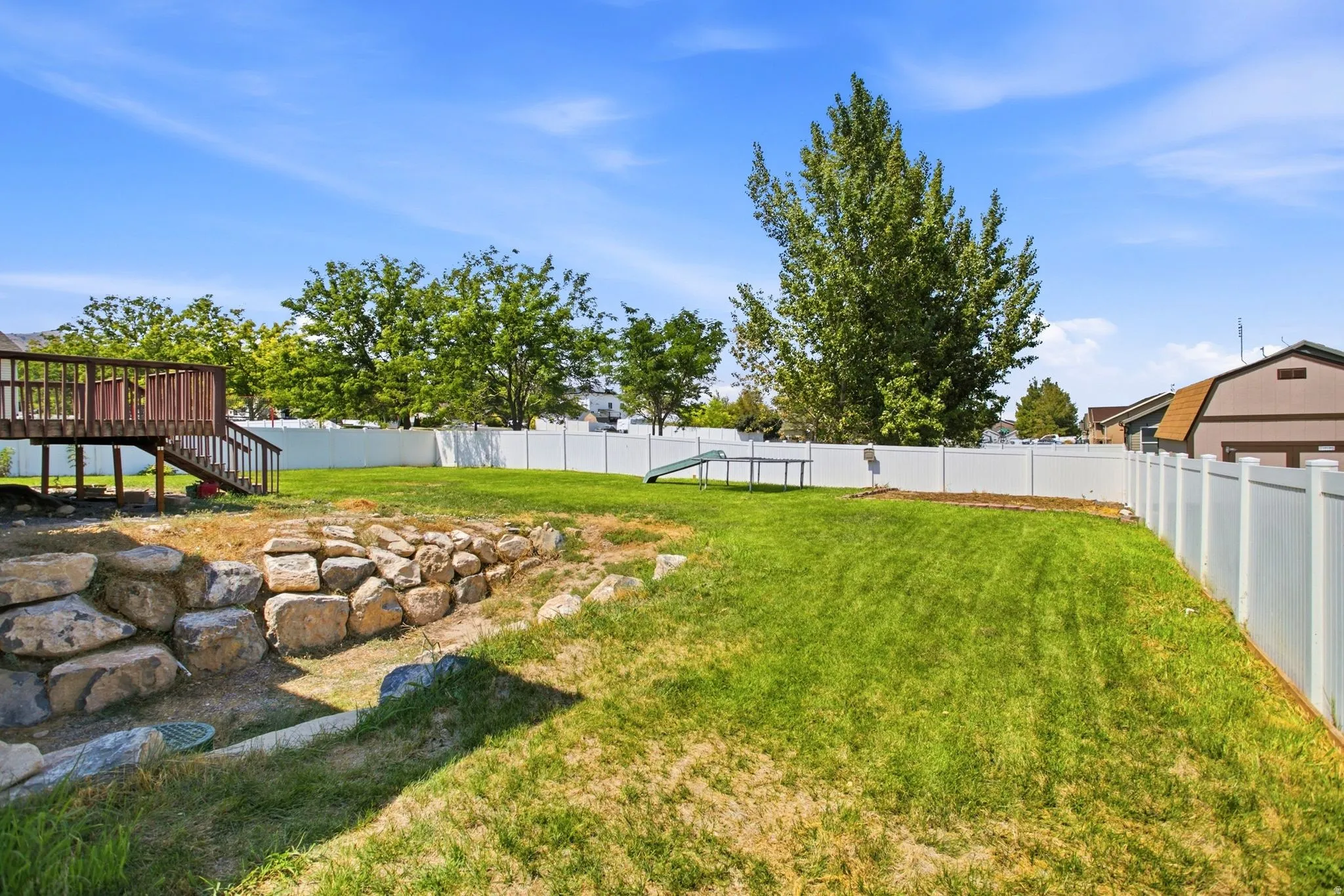 Expansive backyard featuring a wood deck with stairs, a rock retaining wall, a vibrant green lawn, and a white privacy fence
