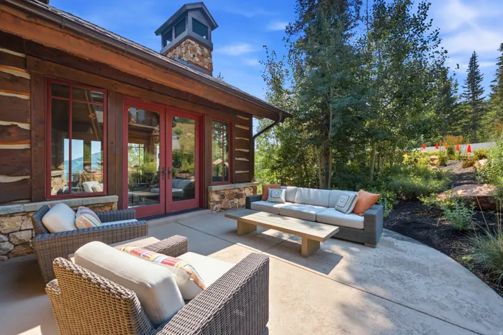 Expansive outdoor patio with concrete flooring, log-style exterior, red-framed double doors, stone chimney, and mature landscaping