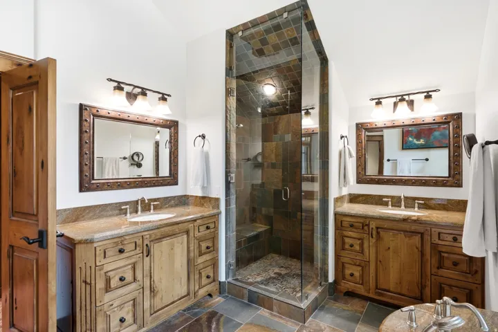 Dual wood-finish vanities with stone countertops, stone tile flooring, and a glass-enclosed shower with stone tile