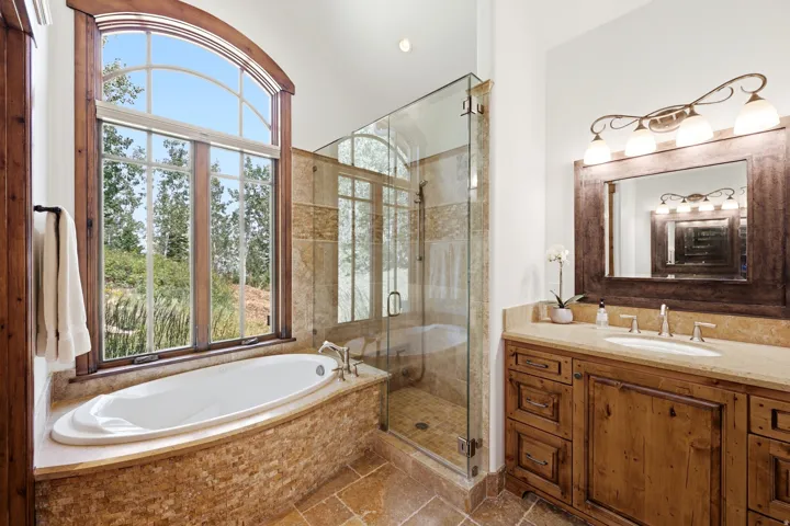Arched window with wood trim, built-in soaking tub, glass-enclosed shower, stone tile flooring, and a wood vanity with a single basin sink