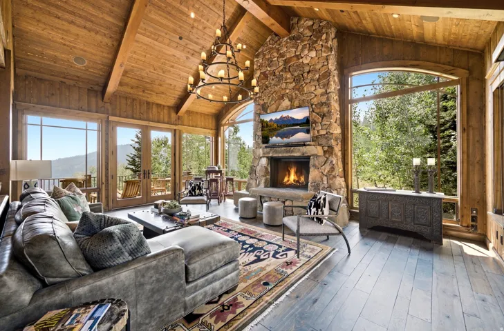 Vaulted wood-paneled ceilings with exposed beams, expansive stone fireplace, large arched window, multiple glass French doors, and wide-plank wood-finish flooring