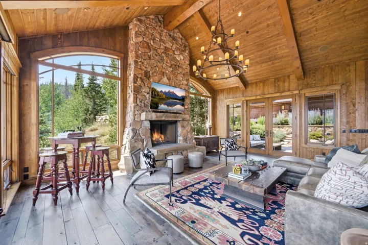 Vaulted wood-paneled ceiling with exposed beams, stone-clad fireplace with hearth, arched window, wood-finish flooring, and double French doors opening to an outdoor area