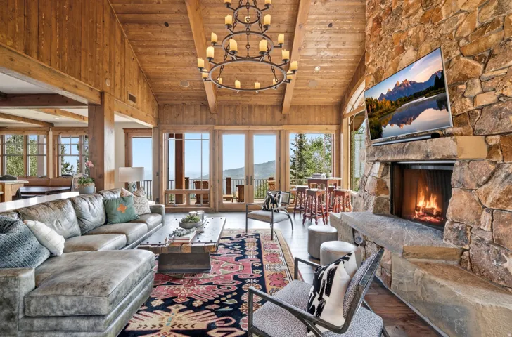 Vaulted wood-paneled ceilings, extensive natural wood accents, a floor-to-ceiling stone fireplace, and multiple glass doors with balcony access