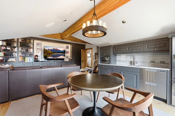 Exposed wood ceiling beams, built-in media center with shelving, recessed lighting, and a full wet bar with paneled cabinetry and integrated stainless-steel appliances