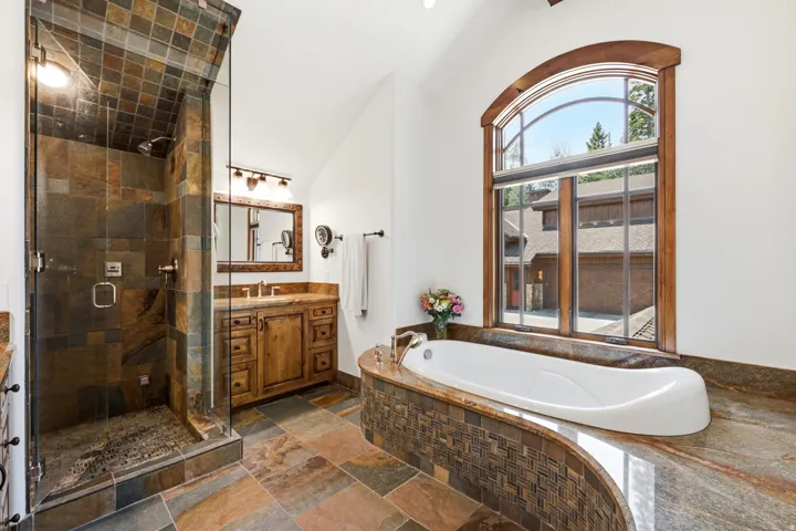 Curved soaking tub with stone surround, walk-in shower with tile walls, wood-finish vanity with countertop, and an arched window
