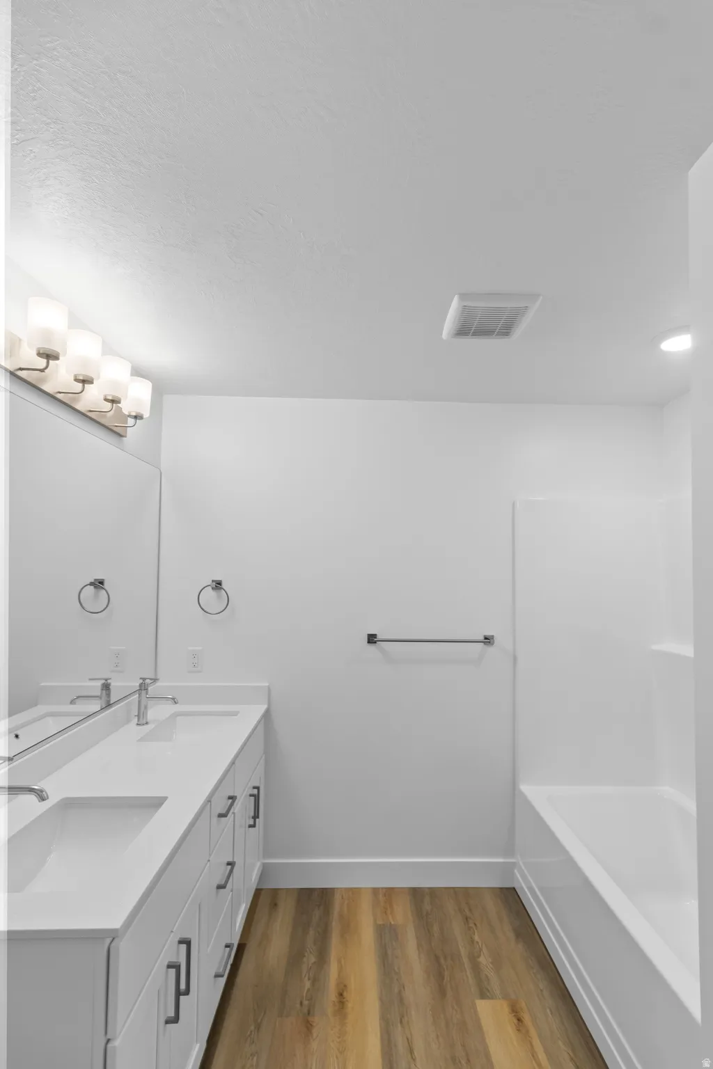Dual vanity with integrated sinks, wood-finish flooring, full-width mirror, and a built-in bathtub