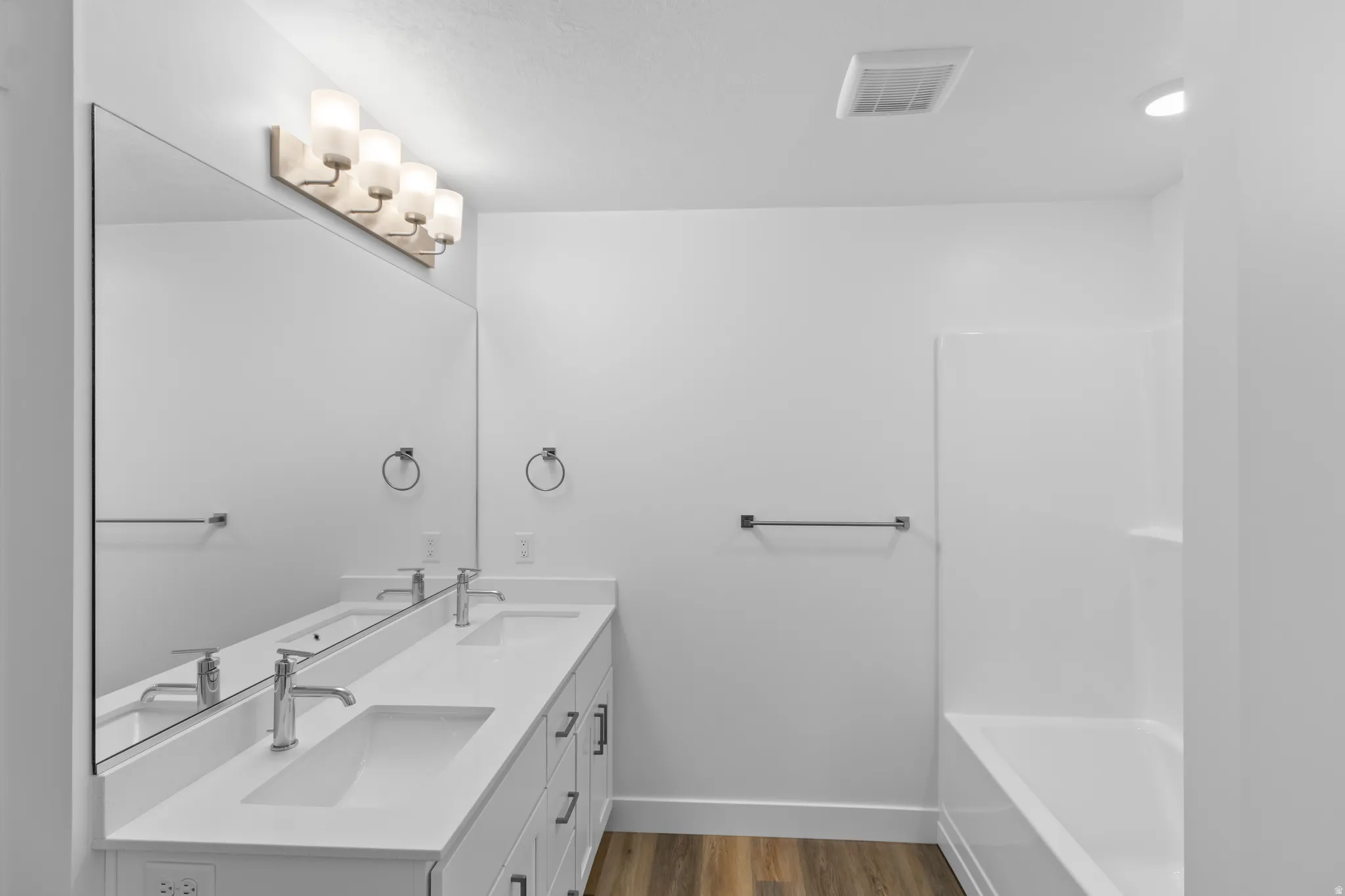 Dual vanity with undermount sinks and chrome fixtures, expansive mirror, wood-finish flooring, built-in bathtub with shower surround
