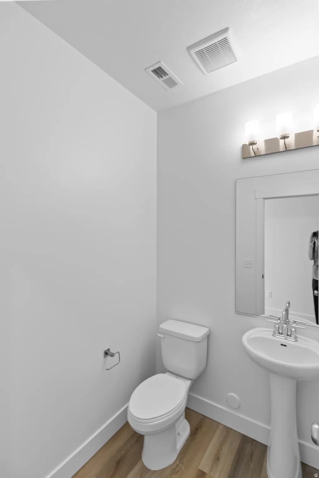 Pedestal sink with chrome-finish faucet, wall-mounted vanity lighting, white toilet, and wood-finish flooring