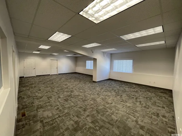 Spacious open room featuring patterned gray carpet, white walls, multiple windows with vertical blinds, two white paneled doors, and a drop ceiling with integrated fluorescent lighting