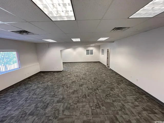 Expansive open-concept space featuring contemporary patterned carpet flooring, white walls, and a suspended acoustic tile ceiling with integrated fluorescent lighting