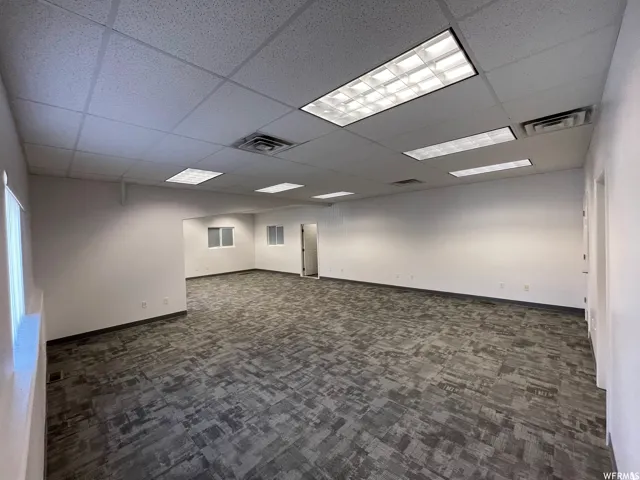 Spacious interior room featuring grey patterned carpet flooring, white painted walls, multiple recessed fluorescent light fixtures, and a suspended acoustic tile ceiling