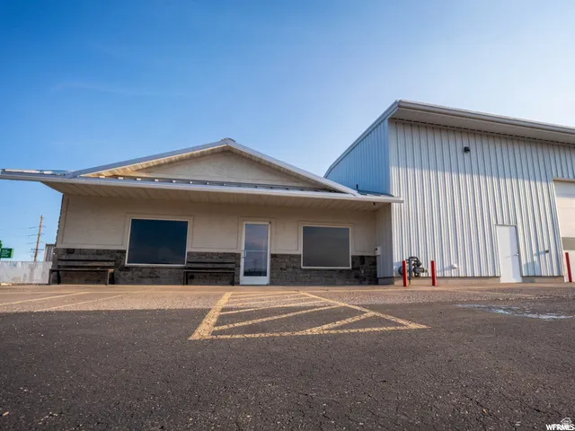 Asphalt parking area with painted lines, stucco exterior building with stone veneer accents, extended awning, single glass entry door, and an adjacent metal-sided structure with a roll-up door