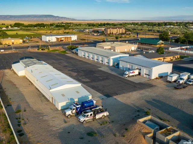Multiple commercial buildings with metal roofs, extensive paved and gravel lot space, and mountain scenery in the background