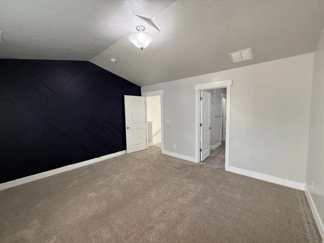 Vaulted ceiling with an attached light fixture, a dark accent wall with geometric paneling, light gray walls, and carpet flooring
