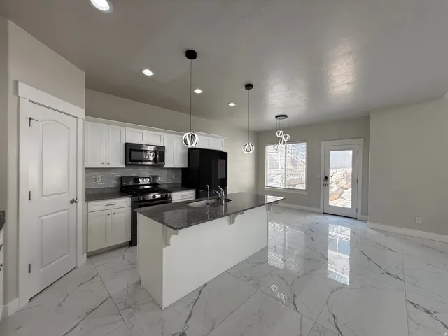 Kitchen featuring white cabinetry, dark stone countertops, stainless steel appliances, a center island with a sink, and pendant lighting