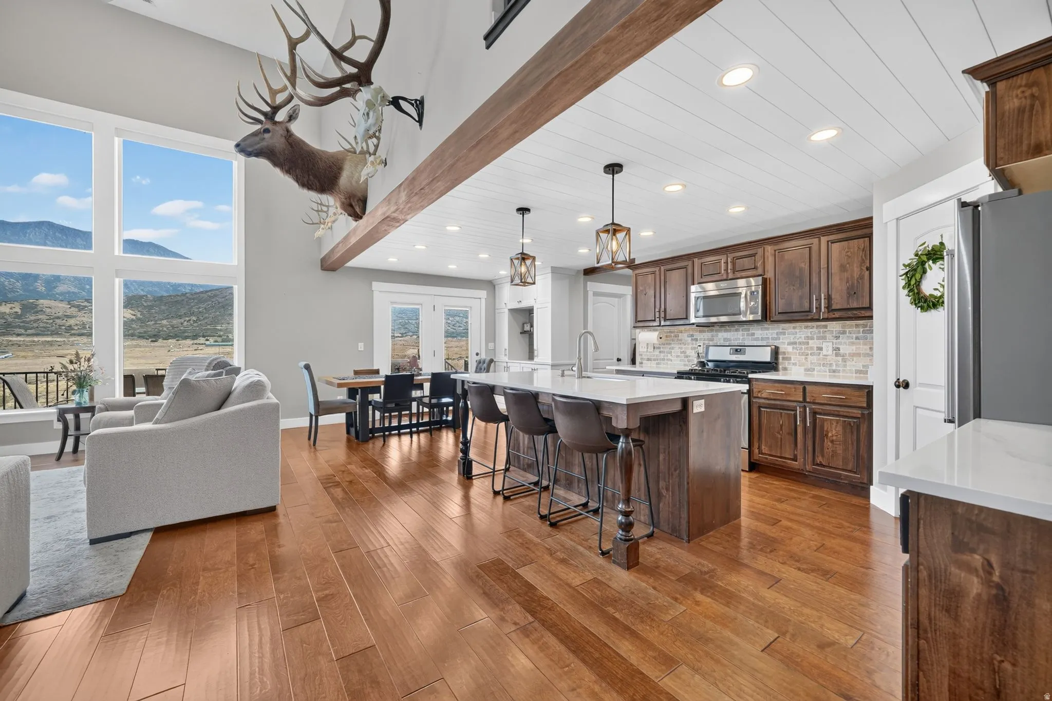 High ceilings with exposed wood beams, expansive floor-to-ceiling windows, wood-finish flooring, a kitchen island with seating, and stainless steel appliances