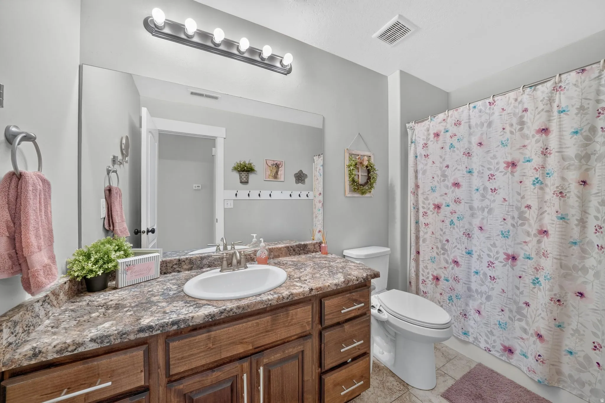 Bathroom featuring a wood-finish vanity with a single basin, a stone-look countertop, a large wall-mounted mirror, and tile flooring