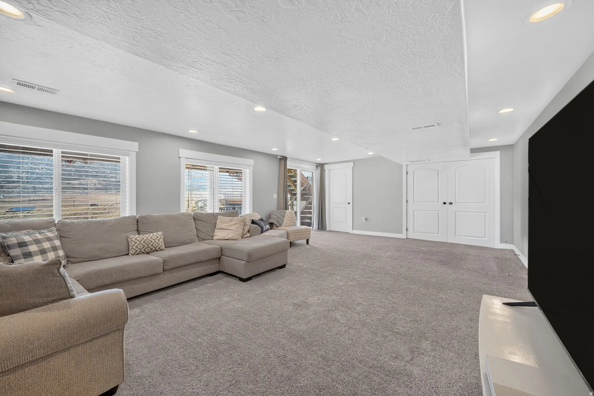 Spacious room featuring light gray walls, recessed lighting, gray carpeting, dual white paneled doors, and two large windows with blinds