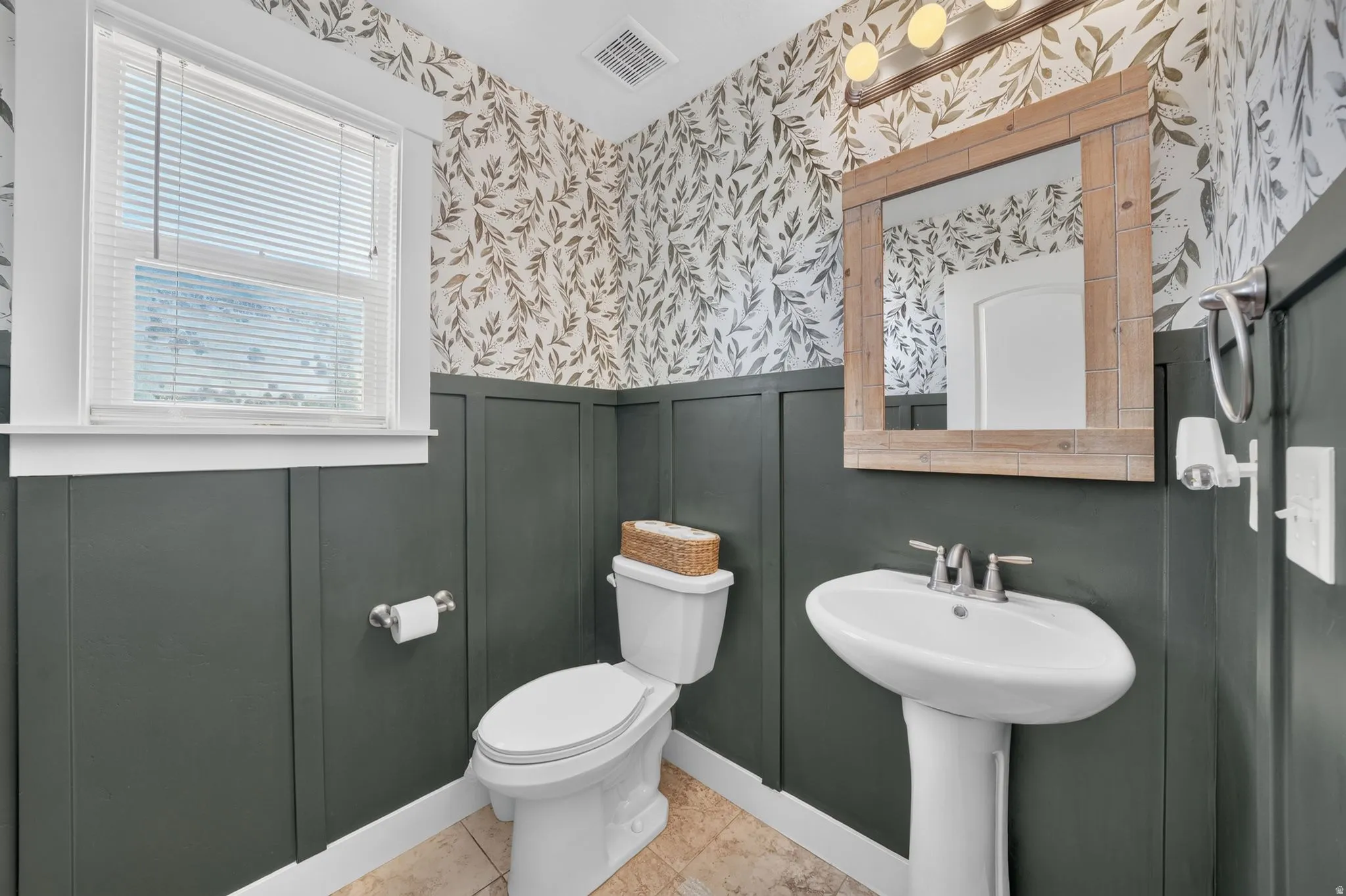 Pedestal sink, wall-mounted mirror with wood-finish frame, contemporary wall-mounted light fixture, decorative wallpaper, and board and batten wainscoting