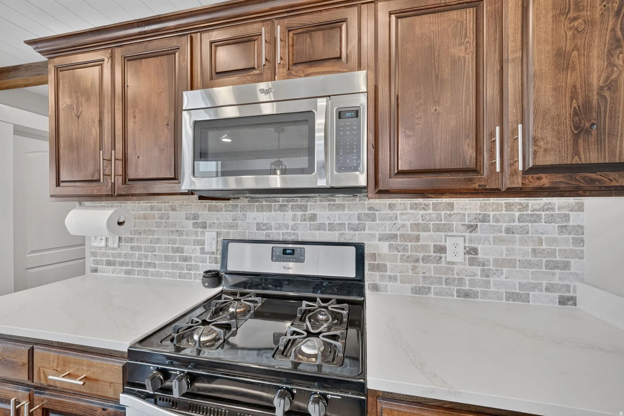 Wood cabinetry with crown molding, stainless steel microwave, stone tile backsplash, white countertops, and a gas range