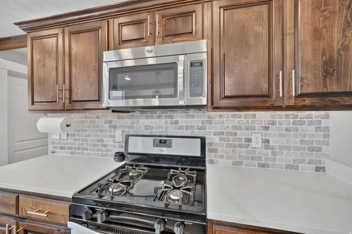 Wood cabinetry with crown molding, stainless steel microwave, stone tile backsplash, white countertops, and a gas range
