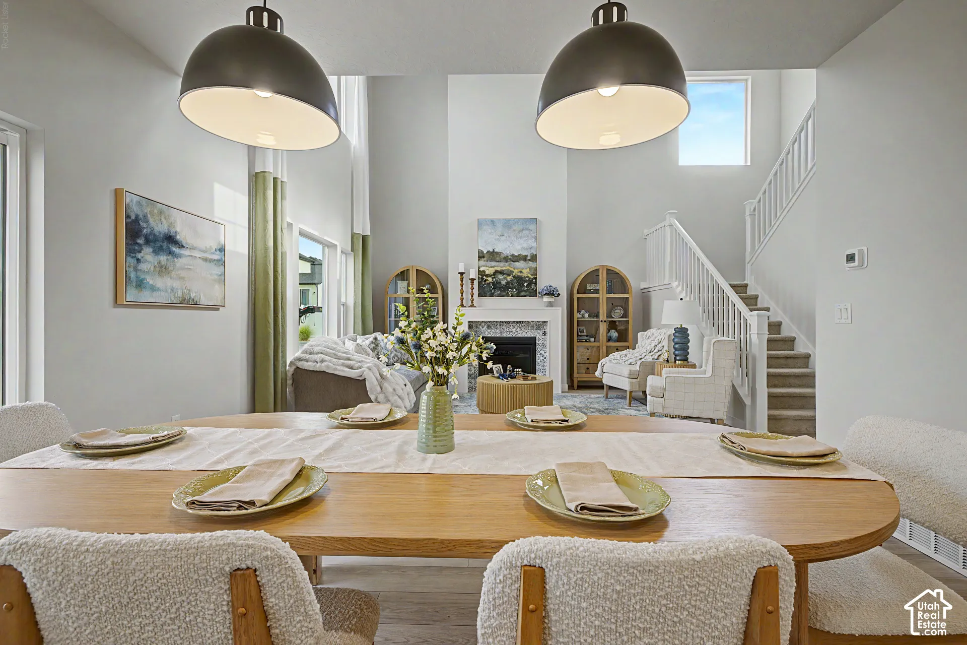 Open-concept living space featuring a wood-finish dining table, double-height ceilings, a fireplace with a stone surround, and a carpeted staircase