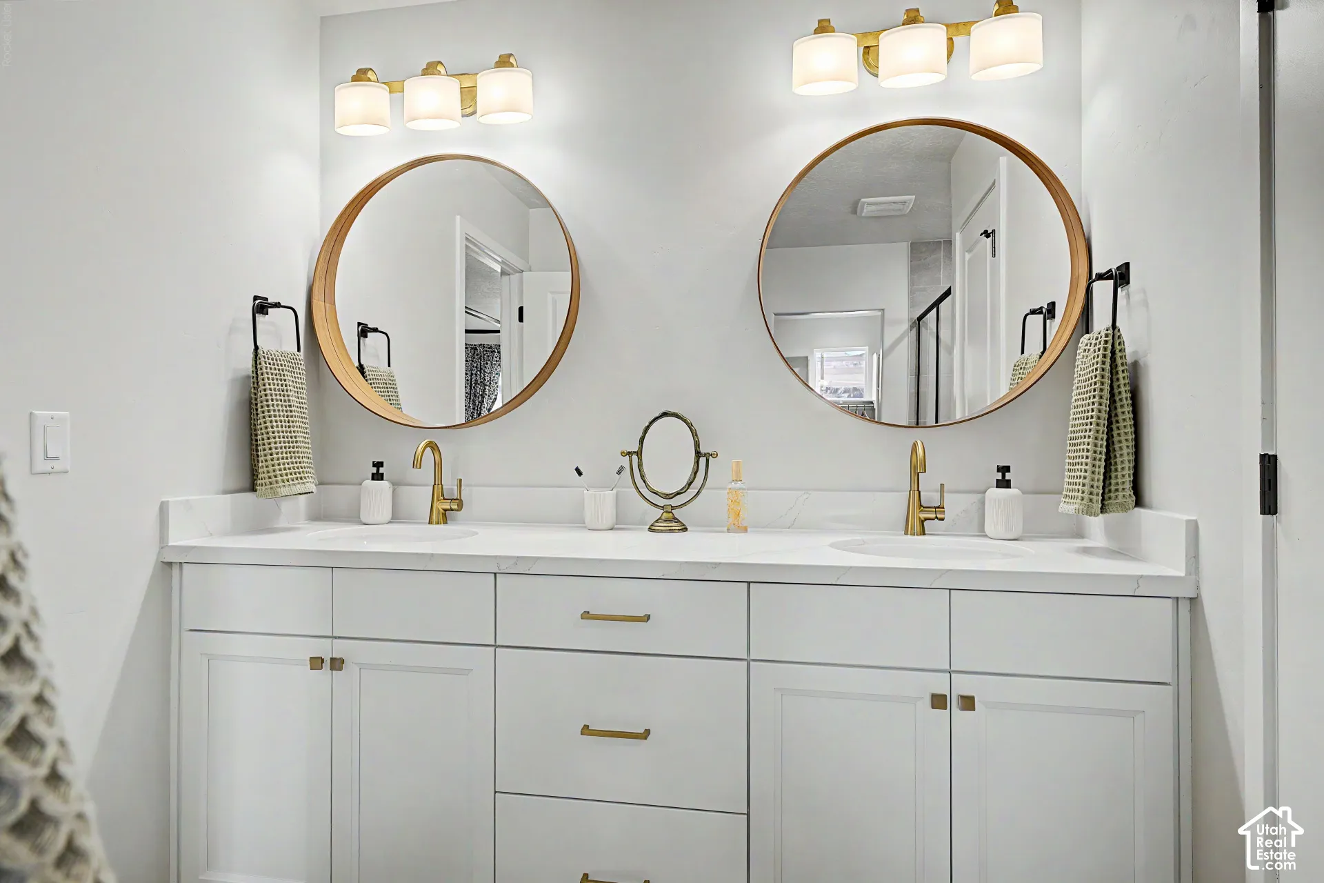 Dual vanity with white cabinetry and gold-tone hardware, white countertop, and two round mirrors, each illuminated by a three-light fixture